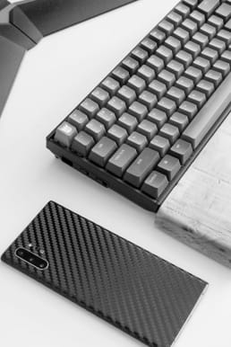Mechanical keyboard and smartphone with carbon fiber case placed on a clean white desk alongside a closed notebook, creating a modern and professional workspace atmosphere